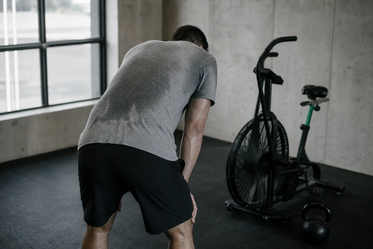 An exerciser bent over with hands on knees after an interval, an air bike behind them